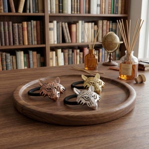 Könnte beinhalten: Ein Holztablett mit drei Haar-Gummis mit Wolfskopf-Motiven in Kupfer, Silber und Gold. Die Haargummis liegen auf einem Holztisch, im Hintergrund ein Bücherregal und dekorative Gegenstände.