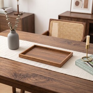 May include: A rectangular wooden serving tray with a smooth, dark brown finish and raised edges. The tray is positioned on a white fabric table runner, centred between a grey ceramic vase containing dried stems and a gold candle holder.