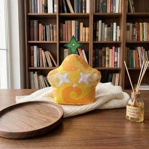 May include: A star-shaped plush toy with a yellow and orange pattern, white star-shaped eyes, and a green star on top. The toy sits on a white knitted blanket, with a wooden tray and a bottle of aromatherapy oil in the foreground.