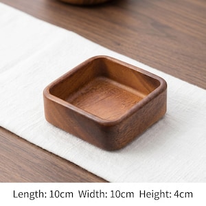 Puede incluir: Un cuenco cuadrado de madera con un acabado de veta marrón natural. El cuenco presenta esquinas redondeadas y bordes lisos. Las dimensiones están claramente indicadas como 10 cm de largo, 10 cm de ancho y 4 cm de alto.