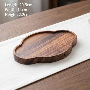 Könnte beinhalten: Ein dunkelbraunes Holztablett mit wolkenartiger Form und glatter Oberfläche. Das Tablett verfügt über eine vertiefte Mitte und erhöhte Ränder. Der Text auf dem Bild gibt Maße von 20,5 cm Länge, 14 cm Breite und 2,2 cm Höhe an.