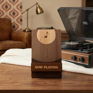 May include: A wooden music-themed phone stand with a musical note and concentric circles design. The stand has the words "NOW PLAYING" engraved on the front. A record player is in the background.