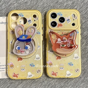 May include: Two yellow phone cases with cartoon character designs. One case features a bunny with a police hat, and the other has a fox. Both cases have a pop-up grip and are decorated with cartoon animal faces. The cases have the text "footopia" and "@zuozan Base".