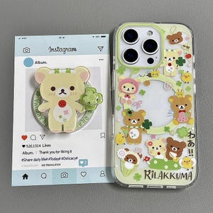 Puede incluir: Una funda transparente para teléfono con un diseño de Rilakkuma, con osos de dibujos animados, tréboles y el texto "Rilakkuma". Al lado, un soporte para teléfono con un diseño de oso y el texto "Rilakkuma".
