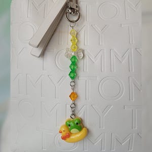 Peut inclure: Un porte-clés perlé avec un fermoir en métal argenté, une perle transparente en forme de nœud et des perles facettées jaunes, vertes et orange. La breloque représente une grenouille verte dans une bouée en forme de canard jaune.