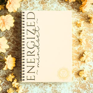 May include: A spiral-bound notebook with the words "ENERGIZED mindset" in black script. The notebook is a light beige color, with a gold-colored chakra symbol at the bottom right. The background is a textured, marbled surface with yellow flowers.