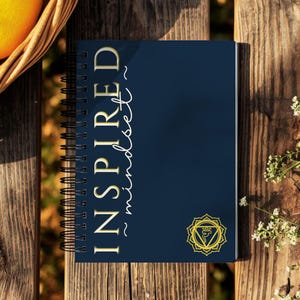 May include: A spiral-bound notebook with a navy blue cover featuring the words "INSPIRED mindset" in gold lettering. A gold-colored symbol is at the bottom right corner. The notebook rests on a wooden surface, with a basket of fruit and flowers nearby.