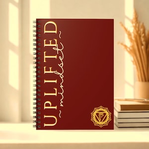 May include: A spiral-bound notebook with a burgundy cover. The words "UPLIFTED mindset" are printed vertically in gold script. A gold chakra symbol is in the lower right corner. The notebook is on a white surface with a vase of dried plants and a stack of books in the background.