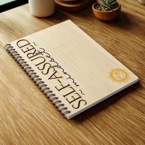 May include: A spiral-bound notebook with a light wood cover. The cover features the words "SELF-ASSURED" in black and gold lettering, along with a gold chakra symbol. The notebook is open on a wooden surface.