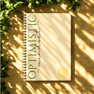 May include: A spiral-bound notebook with the words "OPTIMISTIC mindset" in green script. The notebook has a cream-colored cover with a gold-colored chakra symbol in the lower right corner. Green leaves frame the top and left side.