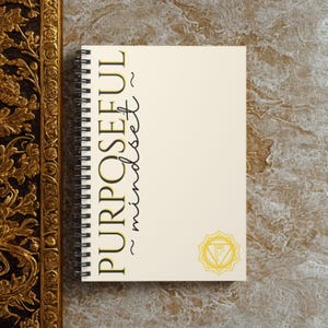 May include: A spiral-bound notebook with the words "PURPOSEFUL mindset" in black and gold lettering. A gold-colored geometric design is in the lower right corner. The notebook is on a textured, neutral-toned surface.