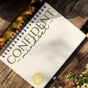 May include: A spiral-bound notebook with the words "CONFIDENT mindset" in gold and black lettering. The notebook is open on a wooden surface, with a yellow geometric design at the bottom. The background is blurred with natural light.