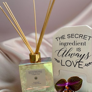 May include: A pearlescent glass reed diffuser with a gold cap and wooden reeds. The label reads "ANITA'S NATURALS" and "Puanani 7 oz Reed Diffuser." A small sign says "THE SECRET ingredient IS Always LOVE." A purple orchid is in the foreground.