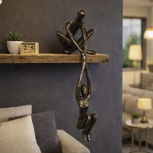 Può includere: Scultura color bronzo raffigurante due figure, una seduta sopra e l'altra appesa sotto, che si tengono per mano. La scultura è montata su una mensola di legno, con una piccola pianta e un calendario. Le figure sono stilizzate.