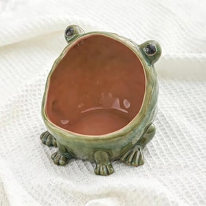 Puede incluir: Un cuenco de cerámica con forma de rana con una amplia abertura y un interior marrón. El exterior es de color verde moteado con ojos negros y patas de rana. El cuenco está diseñado para colocarse sobre una superficie plana.