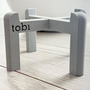 May include: A gray plant stand with the word "tobi" printed on the front. The stand has a cross-shaped design with four legs. The stand is made of a solid material and is designed to hold a plant pot.