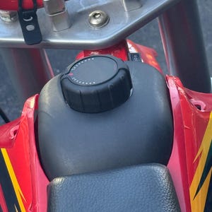 May include: Close-up of a red and black motorcycle. The image shows the handlebars, fuel tank with a black cap, and seat. The motorcycle has yellow and black accents. The handlebars are silver.