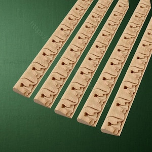 May include: Close-up of several unfinished wooden decorative trim pieces. Each piece features a repeating carved design of stylized leaves and hearts. The wood has a natural, light brown color. The trim pieces are angled across a green background.