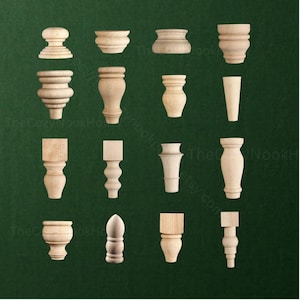 May include: A collection of sixteen unfinished light-colored wooden furniture feet and knobs arranged in a grid. The pieces feature various turned, tapered, and square-topped designs, showcasing natural wood grain textures and smooth, rounded profiles.
