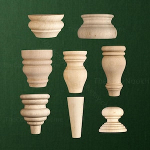 May include: A collection of eight unfinished light-colored wooden furniture feet and knobs in various shapes, including bun feet, tapered legs, and rounded finials, arranged in a grid pattern against a dark green background.