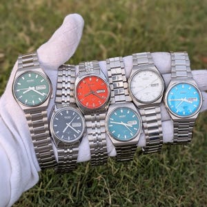 Puede incluir: Una colección de seis relojes de pulsera automáticos Seiko con correas de eslabones de metal plateado. Los relojes cuentan con esferas redondas en verde, negro, naranja, turquesa, blanco y azul claro, cada uno con ventana de día y fecha.
