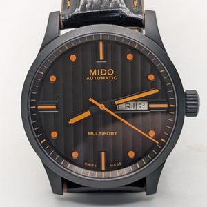 Puede incluir: Reloj automático Mido Multifort negro con esfera negra, detalles en naranja y correa de cuero negra. La esfera muestra el día y la fecha. El reloj tiene el texto "MIDO AUTOMATIC" y "MULTIFORT" en la esfera.