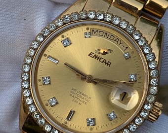 May include: A gold-toned wristwatch with a gold dial and a bezel encrusted with small, sparkling stones. The watch face displays the day of the week, the date, and the brand name "ENICAR". The watch band is also gold-toned.