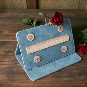 May include: A blue tablet case with a wooden ruler and heart-shaped magnets. The ruler is marked in inches. A red rose and other roses are in the background. The case is open, revealing a second layer.