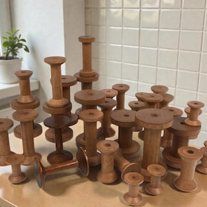 May include: A collection of various sized wooden spools, arranged on a light-colored surface. The spools are cylindrical with a flat top and bottom, and are made of a light brown wood. Some spools are stacked, creating a visually interesting display.