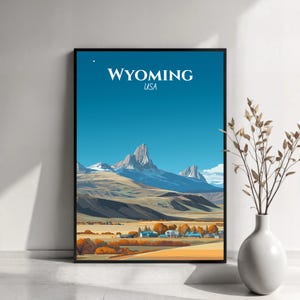 Könnte beinhalten: Ein gerahmter Kunstdruck mit einer stilisierten Landschaft von Wyoming, USA. Das Kunstwerk zeigt eine Bergkette unter einem klaren blauen Himmel, mit dem Wort "WYOMING" und "USA" in weißem Text. Die Szene umfasst ein Tal mit Bäumen und Gebäuden.
