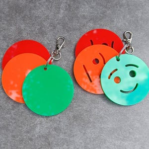 May include: Two sets of circular acrylic keychains attached to silver-toned metal lobster clasps. Each set features three stacked discs in red, orange, and green. One green disc displays a cutout smiley face design, while the others are solid.
