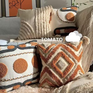 Può includere: Una collezione di cuscini decorativi con vari design e texture. I cuscini presentano colori crema, arancione e grigio con motivi geometrici, dettagli trapuntati e finiture con frange. La parola "TOMATO" è visibile nell'immagine.