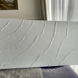 May include: A rectangular, white abstract art piece with a textured surface. The artwork features curved, raised lines across the canvas, creating a wave-like pattern. The piece is displayed indoors, leaning against a wall.