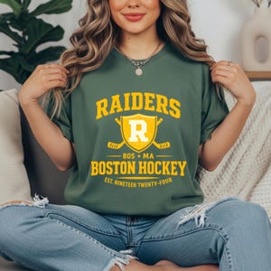 May include: Green t-shirt with the text "RAIDERS BOSTON HOCKEY" in yellow. The design includes a shield with the letter "R" and crossed hockey sticks. The text "EST. NINETEEN TWENTY-FOUR" is also visible.