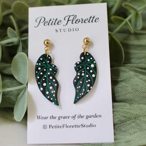Puede incluir: Pendientes con forma de hoja, con fondo negro, vetas verdes y lunares blancos. Cuelgan de postes dorados. La tarjeta detrás de los pendientes dice "Petite Florette Studio" y "Wear the grace of the garden."