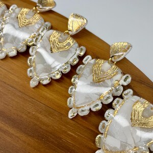 Peut inclure: Gros plan de plusieurs boucles d'oreilles blanches et dorées. Chaque boucle d'oreille a une forme triangulaire avec un centre blanc marbré, une bordure dorée et un accent doré en haut. Les boucles d'oreilles sont présentées sur une surface en bois.