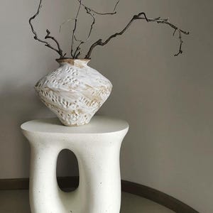 May include: A white, speckled, sculptural side table with a central oval cutout, supporting a textured, off-white ceramic vase with a wide, angular body and a narrow neck, containing several thin, dark brown, leafless, twisted branches.