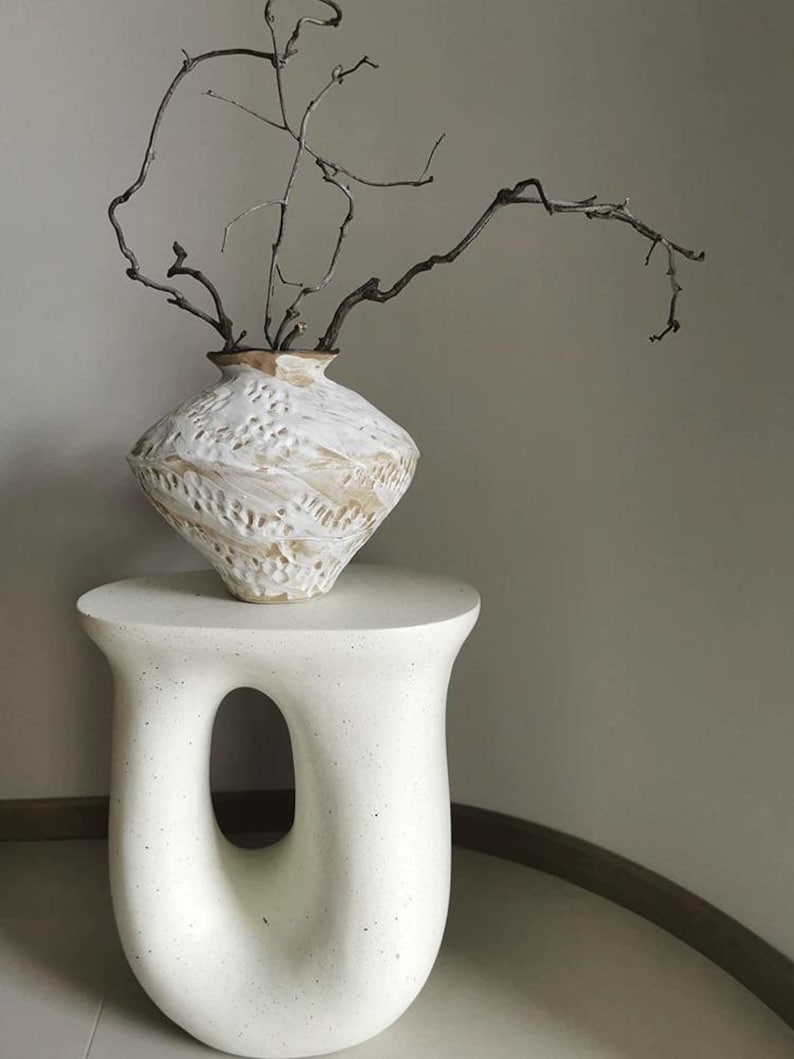 May include: A white, speckled, sculptural side table with a central oval cutout, supporting a textured, off-white ceramic vase with a wide, angular body and a narrow neck, containing several thin, dark brown, leafless, twisted branches.