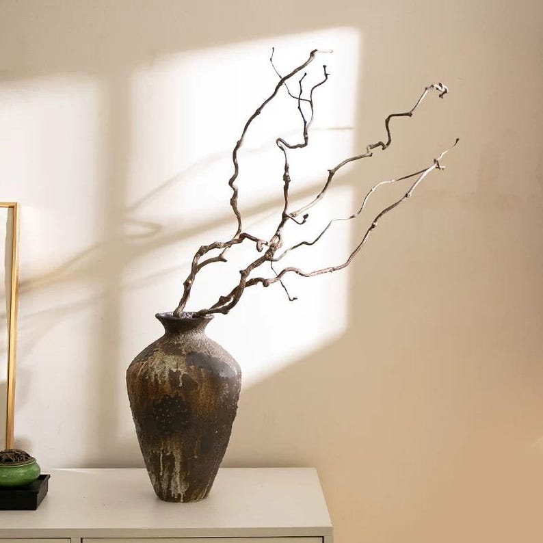 May include: A dark brown, textured ceramic vase with a tapered neck and a wide, rounded body. The vase contains several long, thin, twisted, and gnarled brown branches that extend upward and outward in a branching, organic pattern.