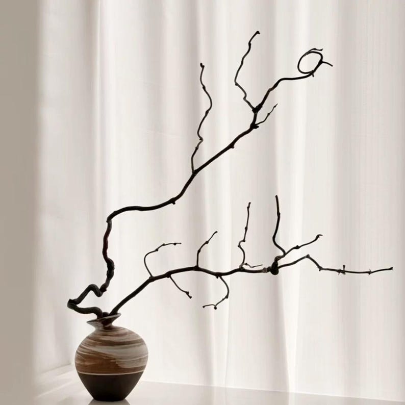 May include: A small, round ceramic vase with a dark brown base and a swirling white and tan marbled pattern on the upper portion. A single, long, thin, dark brown, gnarled branch extends upward and outward from the narrow opening of the vase.