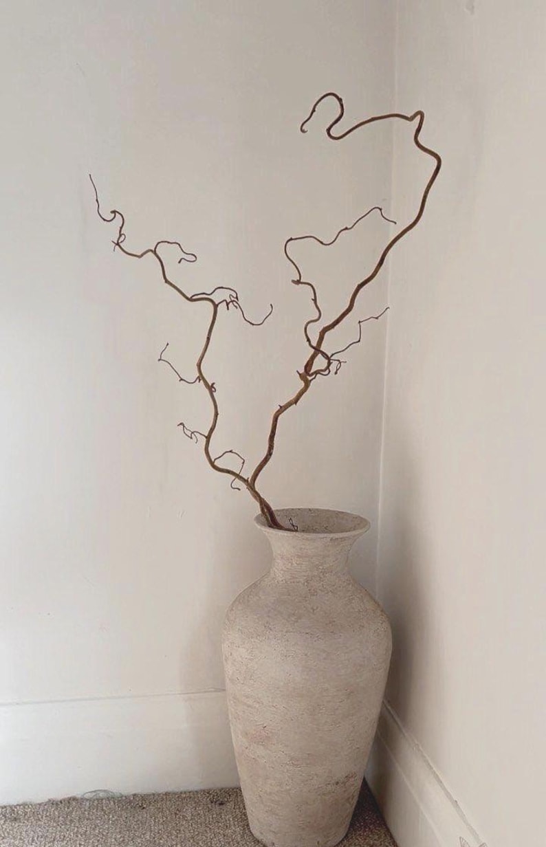 May include: A tall, off-white ceramic vase with a textured, matte finish and a flared rim. A single, long, brown, twisted willow branch with multiple thin, curling stems extends upward from the opening of the vase, creating a natural, organic shape.
