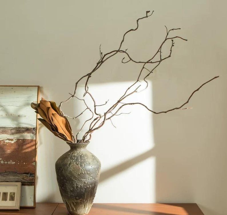 May include: A dark gray, textured ceramic vase with a bulbous body and a narrow neck. The vase contains a cluster of dried, brown magnolia leaves and several long, thin, winding bare branches that extend upward and outward in a natural pattern.