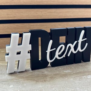 Könnte beinhalten: Ein schwarz-weißes Dekoschild mit dem Text "#text". Das Hashtag-Symbol und das Wort "text" sind in 3D-Design gehalten. Das Schild steht auf einer hellen Oberfläche mit Holzgrund.