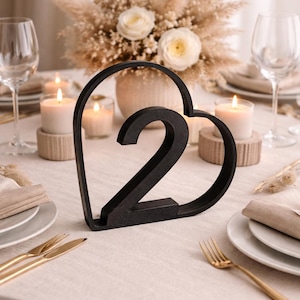 Könnte beinhalten: Schwarze Tischkarte mit der Zahl zwei in Herzform. Der Tisch ist mit weißen Tellern, goldenem Besteck, Kerzen und einem Blumengesteck dekoriert.