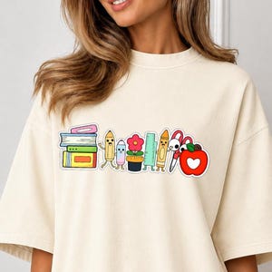 Könnte beinhalten: Ein übergroßes cremefarbenes T-Shirt mit einem Grafikdruck von bunten Schulsachen, darunter ein Stapel Bücher, ein Buntstift, eine Blume im Topf, ein Lineal, ein Bleistift, eine Schere und ein roter Apfel mit einem weißen Herzausschnitt.