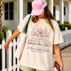 May include: A cream-coloured t-shirt with a design of stacked books tied with a pink bow and floral details. The text reads "A New Chapter Begins" and "Retirement 2026". A pink cap and a green tote bag are also visible in the image.