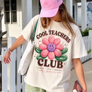 May include: Cream-coloured t-shirt with a large flower design and the text "COOL TEACHERS CLUB". The flower has pink petals and a purple smiley face centre. The shirt also has the text "TEACH + LOVE + INSPIRE". A pink trucker hat is worn.