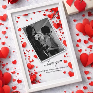 May include: A white framed photo with a black and white image of a couple, surrounded by red paper hearts. The text "I love you" is written on the photo, along with a romantic message. A gift box with a red bow and a red rose are also present.