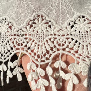 Könnte beinhalten: Nahaufnahme eines weißen Spitzenstoffs mit aufwendigen floralen und geometrischen Mustern. Die Spitze hat einen gewellten Rand mit zarten blattartigen Details. Der Stoff scheint aus Baumwolle oder Ähnlichem zu bestehen, mit einer leicht strukturierten Oberfläche.