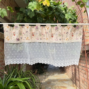 Könnte beinhalten: Ein Scheibengardinen-Vorhang mit einem beigen Oberteil mit buntem Blumenmuster, einem horizontalen Streifen aus cremefarbener Spitze und einem transparenten weißen Unterteil mit Punktmuster und einer gewellten Spitzenkante an einer weißen Stange.